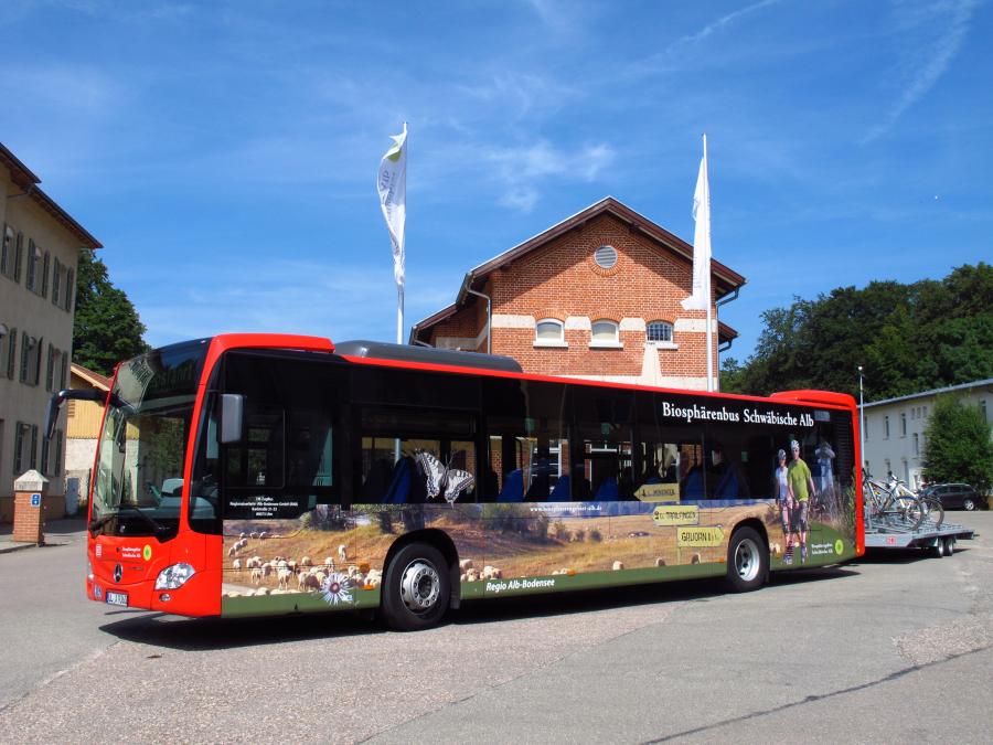 Ein rot-schwarzer „Biosphärenbus Schwäbische Alb“ mit Naturmotiven und Fahrradanhänger steht vor einem Backsteingebäude.