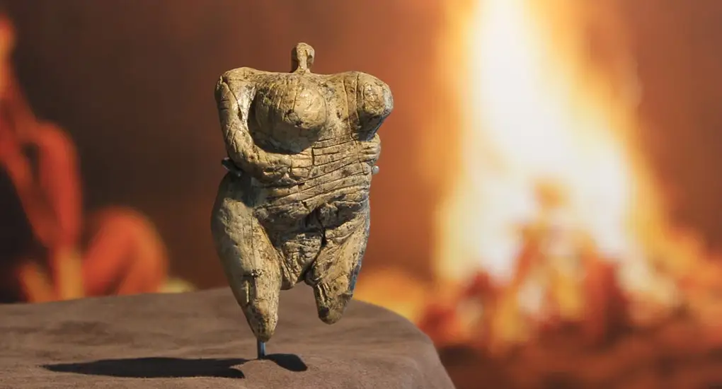 Eine kleine, kopflose Figur aus Elfenbein mit stark betonten weiblichen Rundungen steht auf einem Podest in einem Museum. Die prähistorische Skulptur, bekannt als die Venus vom Hohle Fels, ist vor einem unscharfen Hintergrund mit warmen, rötlich-gelben Lichttönen ausgestellt.