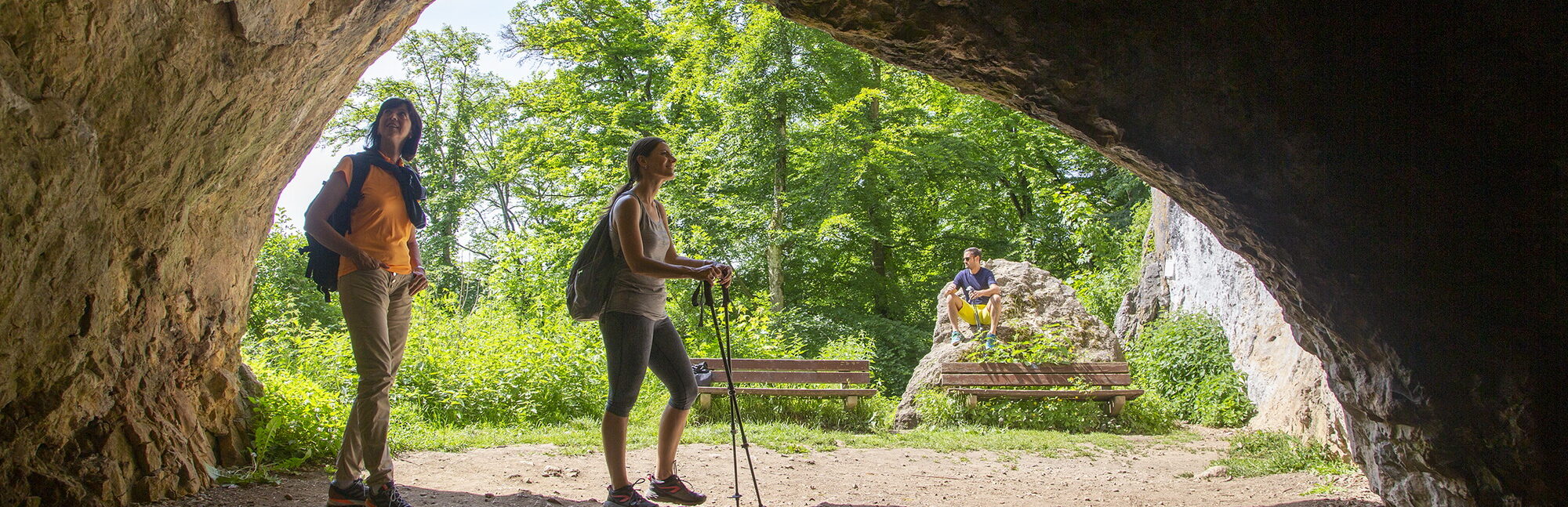 Das Bild zeigt eine Höhle, die einen Blick nach draußen in eine grüne Landschaft ermöglicht. Im Vordergrund sind zwei Wanderer zu sehen: eine Person mit einem Rucksack und eine andere mit Wanderstöcken, die in die Höhle hineinblickt. Im Hintergrund ist eine idyllische Szenerie mit Bäumen und einem Bereich mit Bänken, auf denen eine weitere Person sitzt und die Natur genießt.