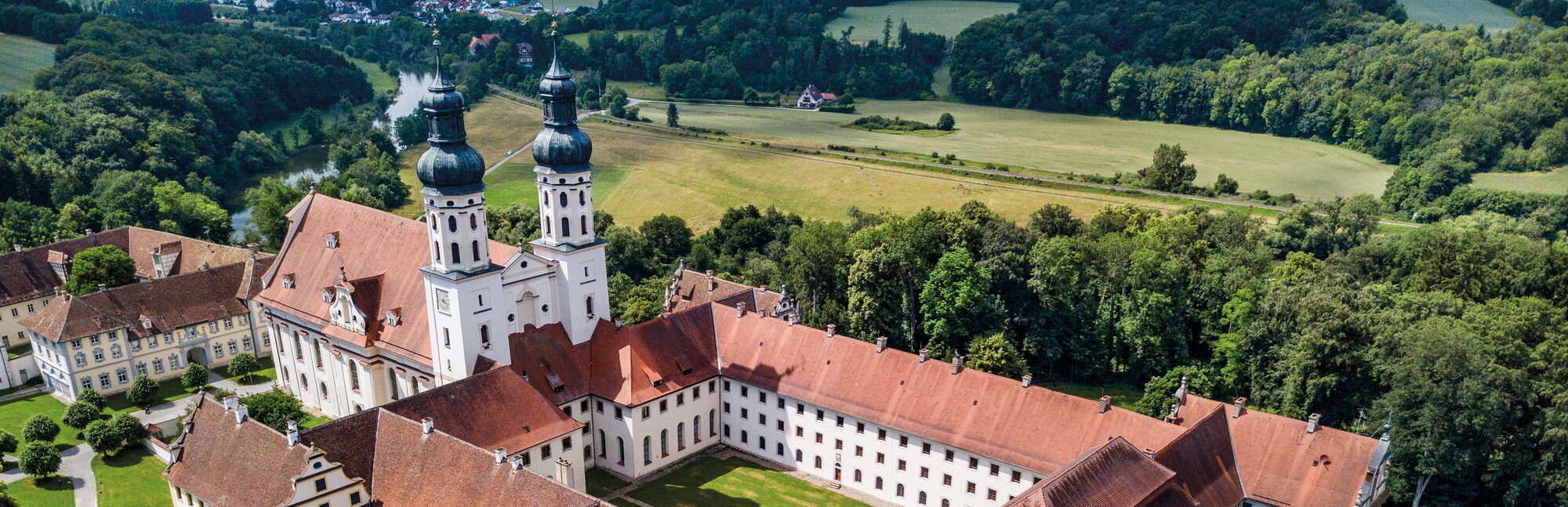 Eine Luftaufnahme vom Kloster Obermarchtal zeigt ein beeindruckendes Klostergebäude mit roten Ziegeldächern und markanten Türmen, das von weitläufigen Grünflächen und Bäumen umgeben ist und eine harmonische Verbindung zwischen Architektur und Natur vermittelt.