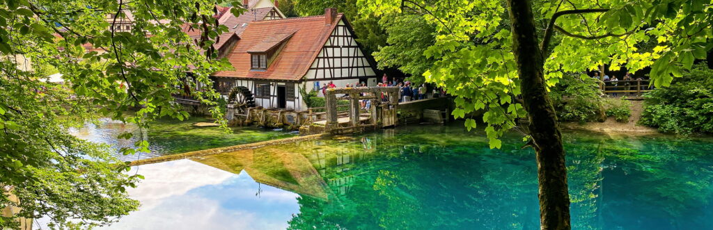 Ein malerischer Blick auf ein traditionelles Fachwerkhaus am Blautopf, blauen Sees, umgeben von üppigem Grün und Bäumen.