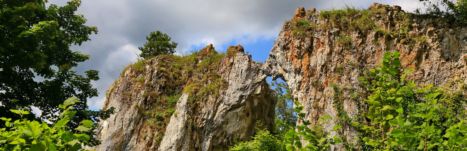 Eine imposante helle Felswand ragt steil aus einem dichten, grünen Wald empor. Der Fels ist von Bäumen umgeben, die bis zum Rand des Felsens wachsen. Rechts davon führt ein geschwungener Weg durch eine sonnige Wiesenlandschaft mit vereinzelten Bäumen und einem kleinen Fluss, der sich durch die Wiesen schlängelt. Im Hintergrund erstreckt sich ein bewaldeter Hügel unter einem blauen Himmel mit leichten Schleierwolken.